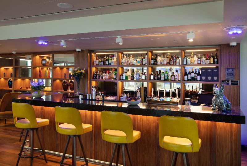 Tower Suites By Blue Orchid London Kültér fotó The photo shows a modern bar area. In the foreground, there are several yellow bar stools lined up in front of a sleek black countertop. Behind the bar, there is a well-stocked shelf displaying a variety of bottles, including spirits and liqueurs, il
