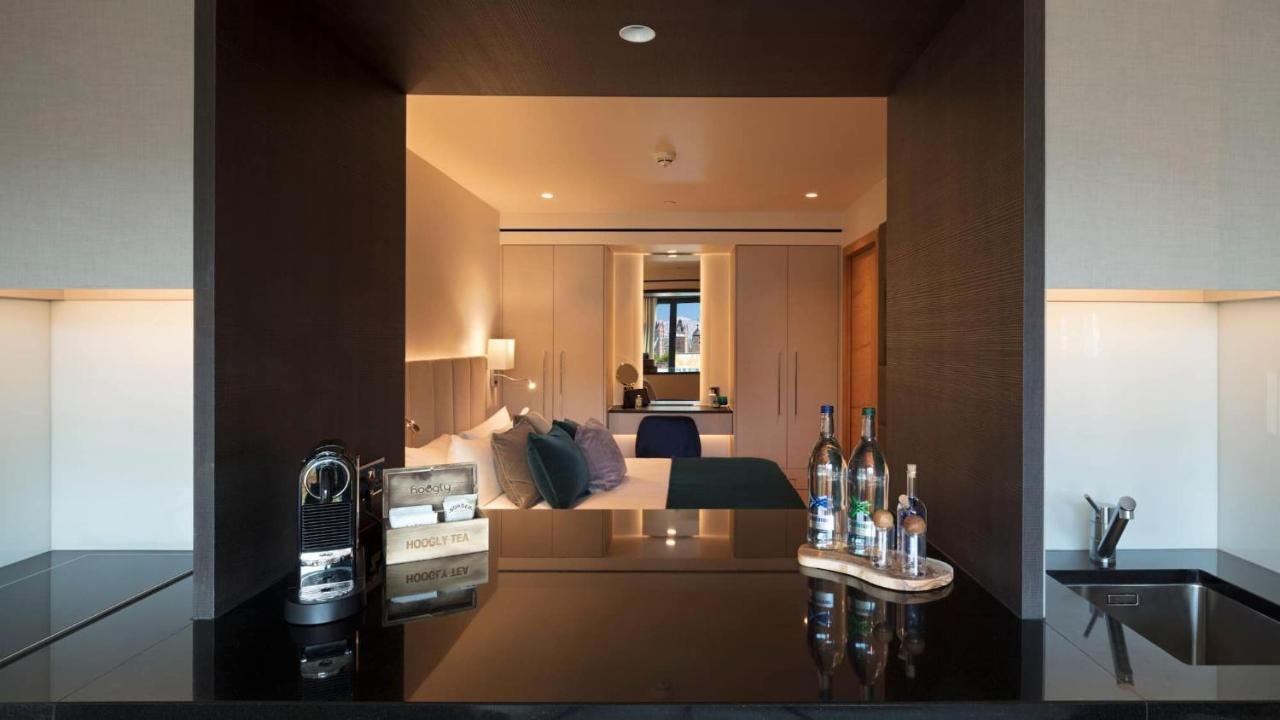 Tower Suites By Blue Orchid London Kültér fotó The image shows a modern hotel room with a stylish design. In the foreground, there is a dark countertop with several bottles displayed, likely including spirits and water. Just beyond the countertop, you can see the bedroom area, which has light-col