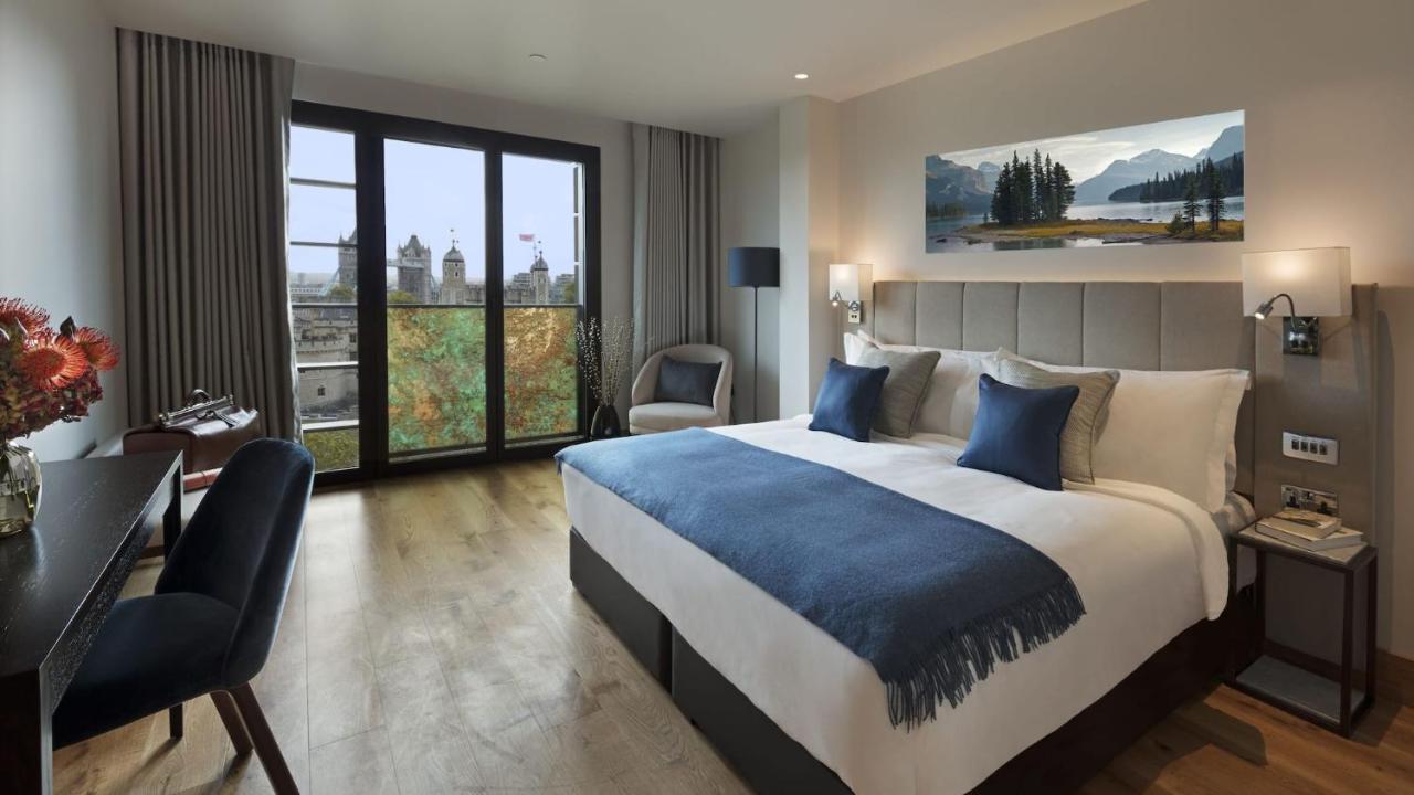 Tower Suites By Blue Orchid London Kültér fotó The photo shows a modern hotel room that features a large bed with white linens and blue accent pillows. There’s a cozy throw blanket at the foot of the bed. The room is well-lit and includes a seating area with a chair and a small table. A large win