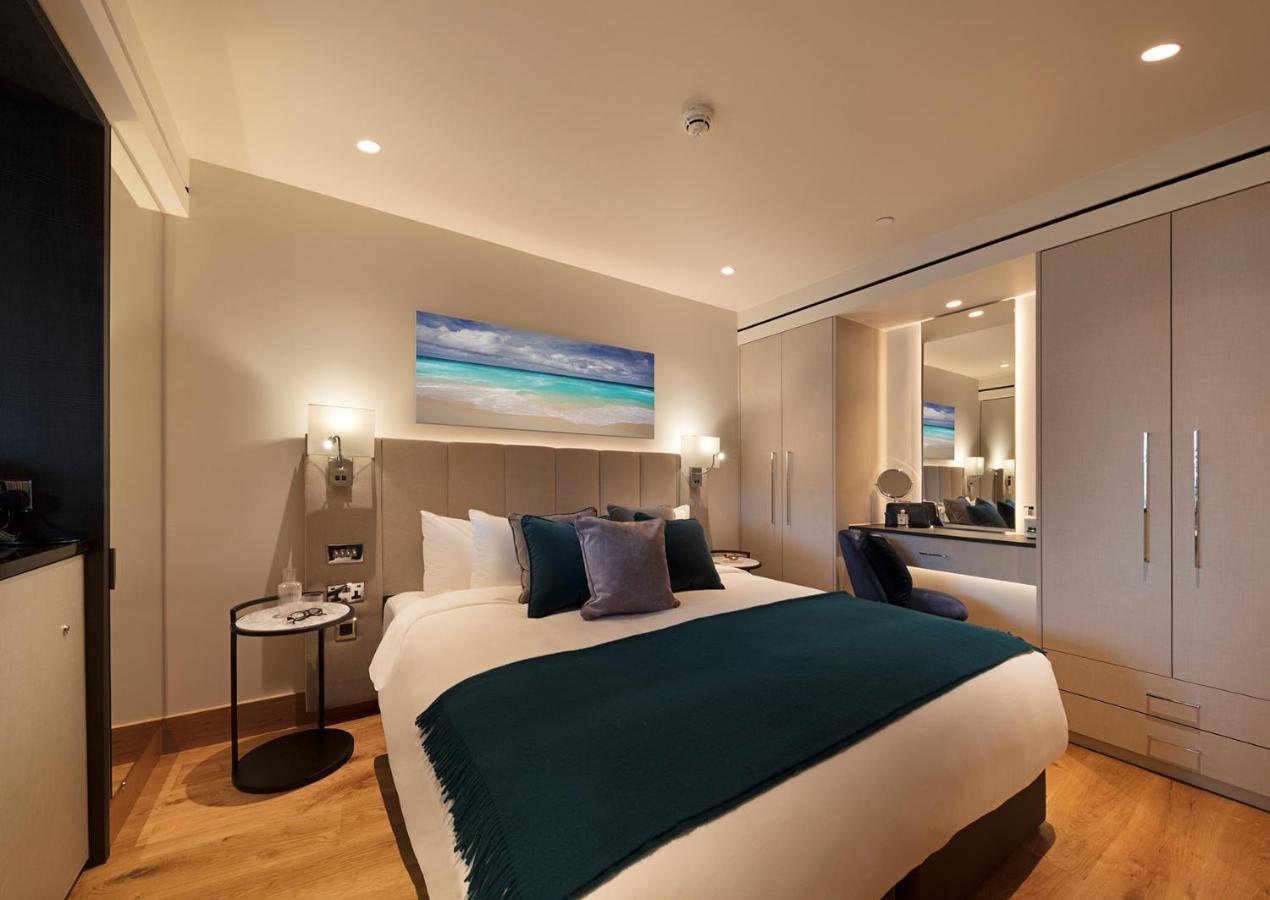 Tower Suites By Blue Orchid London Kültér fotó The photo shows a modern hotel room featuring a double bed with a white duvet and a dark teal throw blanket. There are decorative pillows in shades of blue and purple on the bed. To one side, there's a round bedside table with a small lamp. A large m