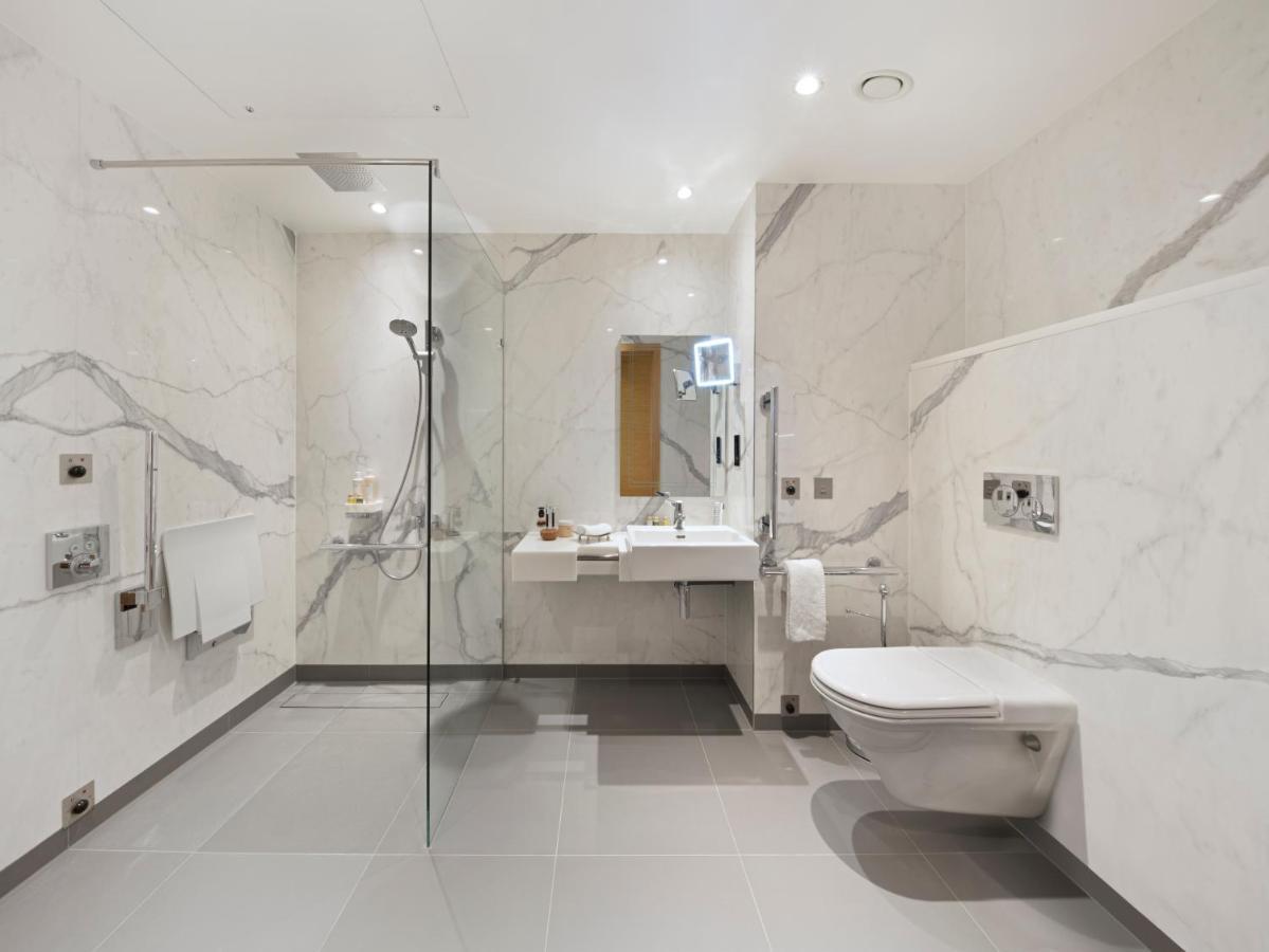 Tower Suites By Blue Orchid London Kültér fotó The photo shows a modern bathroom that features marble walls with a light color. There is a large walk-in shower with a glass partition, and the shower area has a rain shower head. To the right, there is a stylish sink with a mirror above it, and var