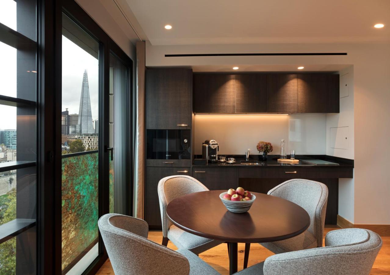Tower Suites By Blue Orchid London Kültér fotó The photo shows a modern kitchen and dining area. It features a round table with four gray chairs, and a bowl of apples is placed in the center of the table. The kitchen has dark cabinetry, a black countertop, and built-in appliances. Large windows a