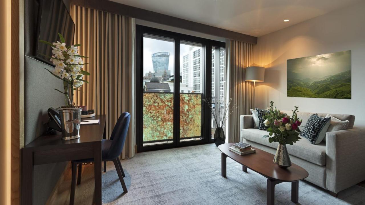 Tower Suites By Blue Orchid London Kültér fotó The photo shows a modern and well-lit living space. There is a comfortable sofa with decorative cushions, a small coffee table in front of it, and a floor lamp providing additional lighting. Beside the sofa, there are decorative plants and a vase wit