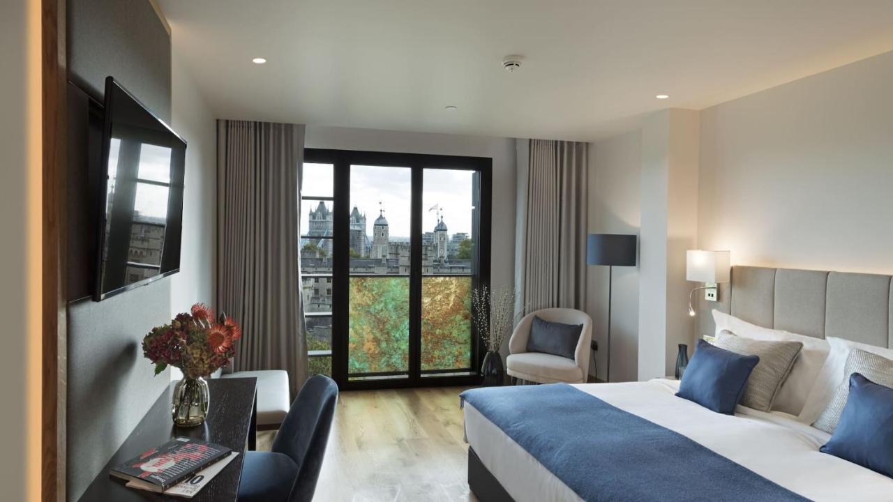 Tower Suites By Blue Orchid London Kültér fotó The photo shows a modern hotel room featuring a large bed with blue and white bedding, a cozy armchair, and a small desk. There is a flat-screen television mounted on the wall across from the bed. The room has large glass doors leading to a balcony o