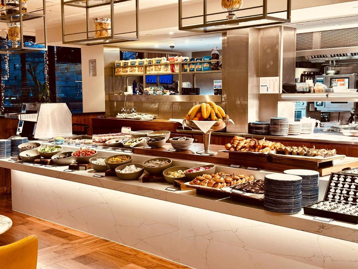 Tower Suites By Blue Orchid London Kültér fotó The photo depicts a buffet-style setup, likely in a restaurant or a hotel dining area. In the foreground, there are multiple bowls and platters filled with a variety of food items, including salads, fruits, pastries, and other dishes. There is a long