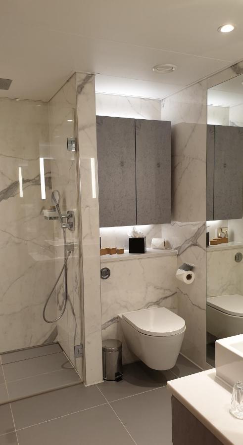 Tower Suites By Blue Orchid London Kültér fotó The photo shows a modern bathroom design featuring elegant marble walls. On one side, there is a transparent shower enclosure with a showerhead and controls visible. Opposite the shower, there is a toilet, and above it are some sleek cabinets. The sp