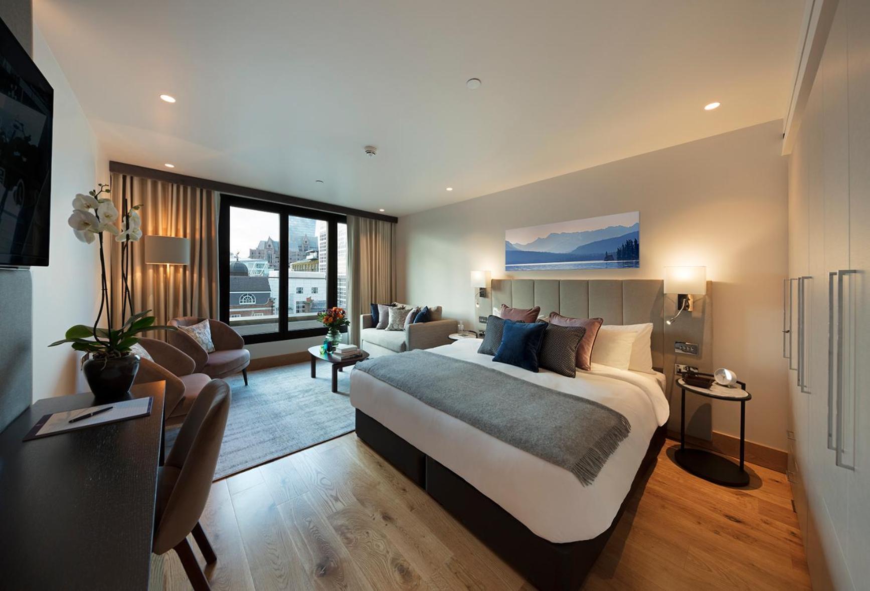 Tower Suites By Blue Orchid London Kültér fotó The photo shows a modern hotel room. In the center, there is a large bed with stylish bedding and pillows in various shades. To one side, there are two comfortable chairs and a small table, creating a cozy seating area. A large window offers a view o